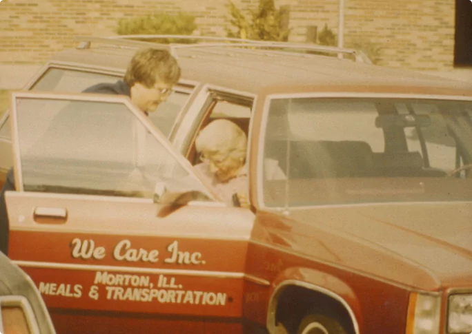 An old photograph of a We Care member helping an elderly woman get into a car